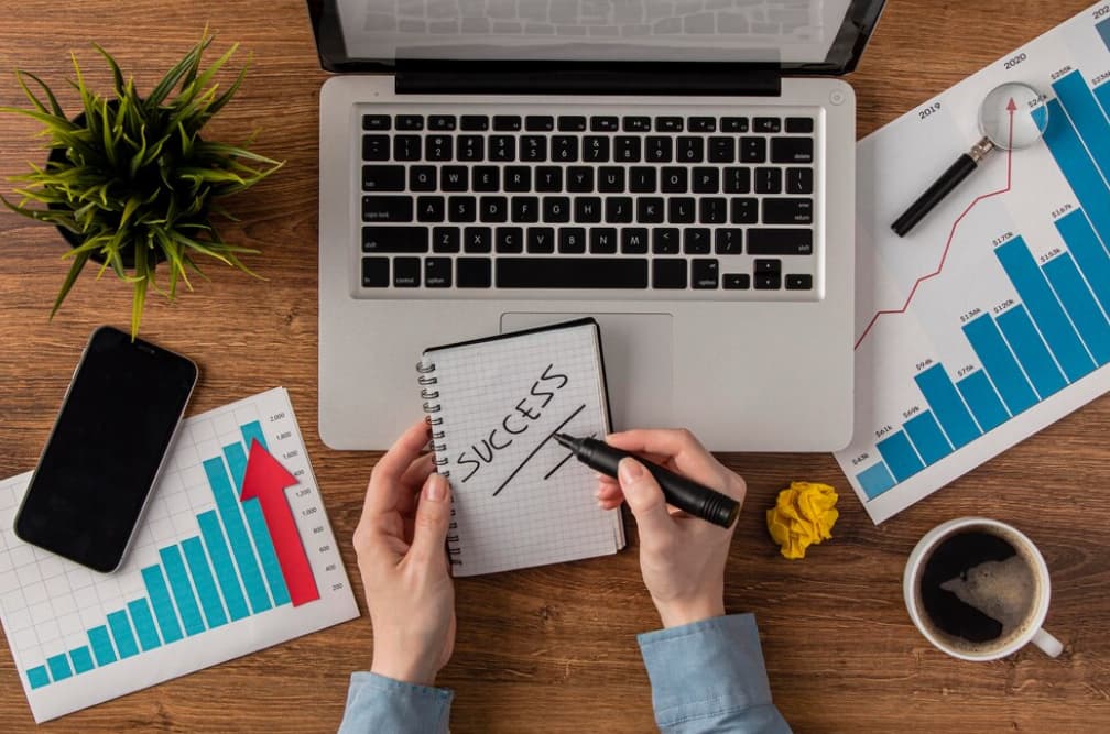 A person writing "SUCCESS" in a notebook at a desk with charts, a laptop, and a coffee cup